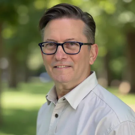 Middle-aged man with glasses wearing a light shirt standing outdoors with blurred green background.