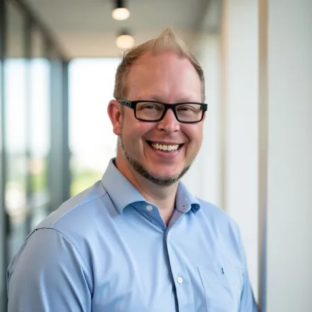 Smiling man with glasses and blue shirt standing indoors near a window with blurred background.