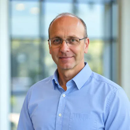 Portrait of a middle-aged man wearing glasses and a light blue shirt with a blurred office background.