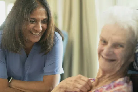 Smiling caregiver in blue scrubs interacts warmly with elderly woman in a cozy indoor setting.