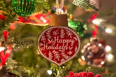 Close-up of a red holiday ornament with Happy Holidays text hanging on a decorated Christmas tree with lights.