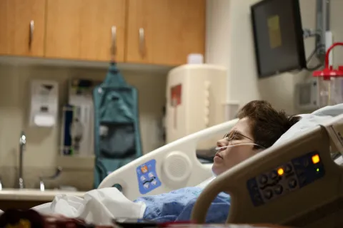 Patient lying in hospital bed receiving oxygen with medical equipment and cabinets in background.
