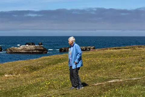 Elderly woman with cane standing on grassy seaside cliff under clear sky near ocean rocks.