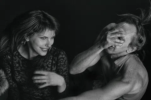 Black and white photo of smiling woman and man covering his face with his hand in dramatic lighting.