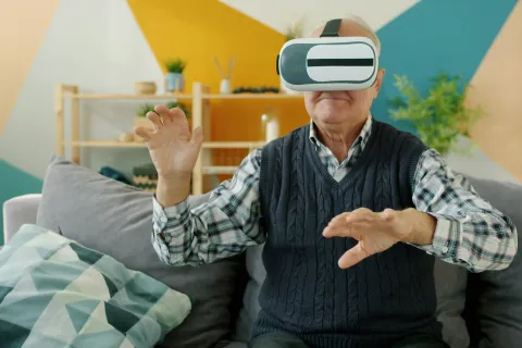 Elderly man wearing virtual reality headset interacting while sitting on a couch indoors with colorful background.