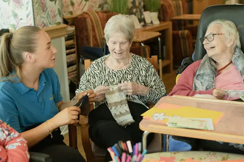 assisted living in atlanta: Young caregiver and two elderly women knitting together and smiling in a cozy room with art supplies on the table.