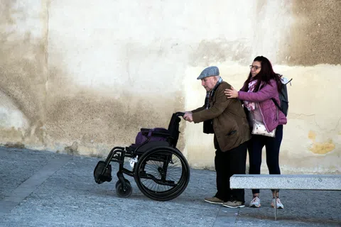 Young woman helping elderly man walk with wheelchair outside against textured wall