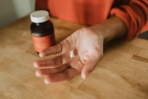 Hand holding a clear probiotic capsule with a bottle labeled probiotic supplement on a wooden table