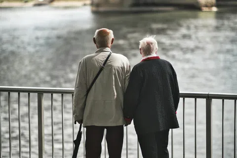 Elderly couple holding hands by a riverside railing under a large bridge on a cloudy day.