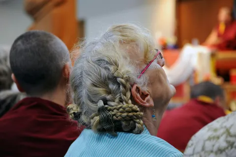 Elderly woman with braided gray hair and pink glasses listening intently during a gathering with others.