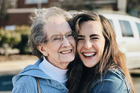 Smiling elderly woman and young woman hugging outdoors, both wearing sweaters and jackets in daylight.