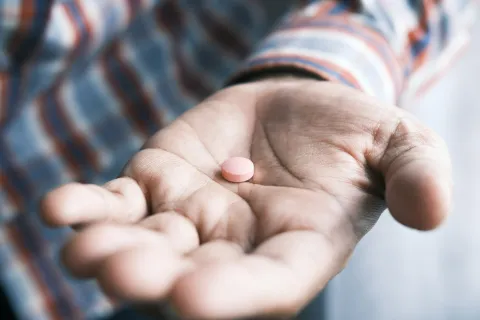 Close-up of an open hand holding a single pink pill with a blurred background of a striped shirt.