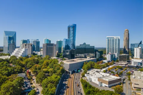 Aerial view of a modern city skyline with skyscrapers, busy highway, and lush green trees under clear blue sky.