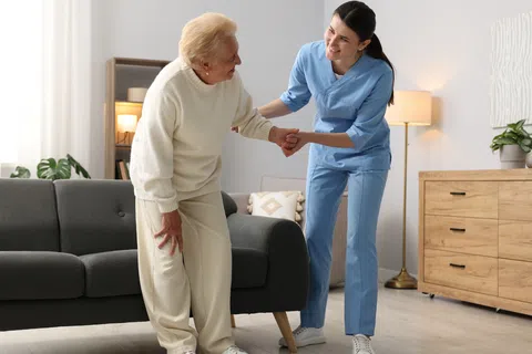 Nurse assisting elderly woman to stand from sofa in a bright, cozy living room with wooden furniture