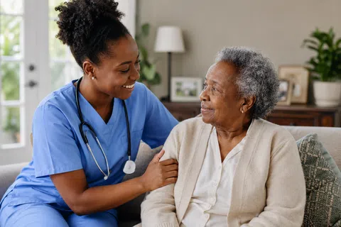 Smiling female nurse in blue scrubs comforting elderly woman sitting on sofa in cozy living room.
