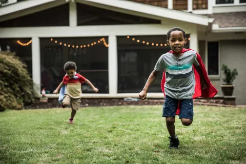 Two young boys wearing red capes running and playing on green grass in a backyard with house in the background