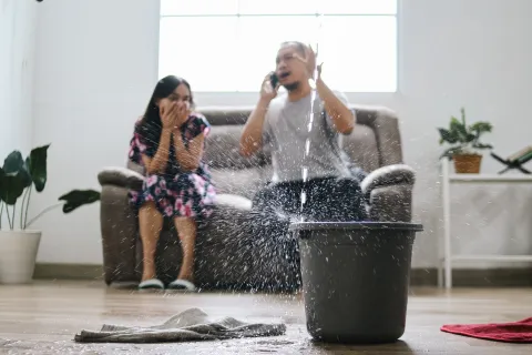Water bursts from overflowing bucket as shocked man and woman sit on couch in bright living room