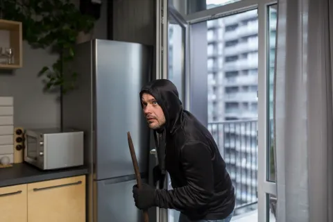 Man in black hoodie and gloves sneaking inside a modern kitchen holding a crowbar, near an open balcony door.