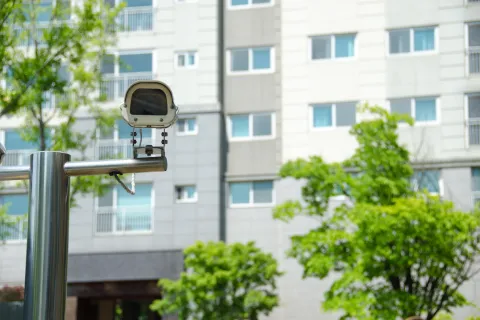 Security camera mounted on metal pole outside apartment building surrounded by green trees under sunlight.