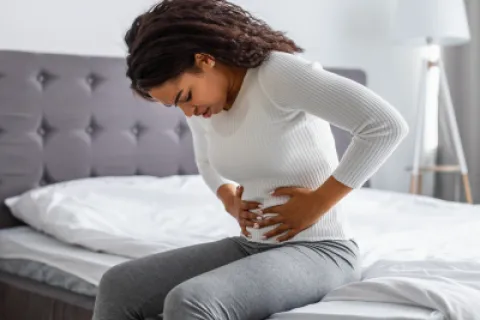 Woman sitting on bed clutching her stomach in pain, wearing white sweater and gray pants in bright bedroom.