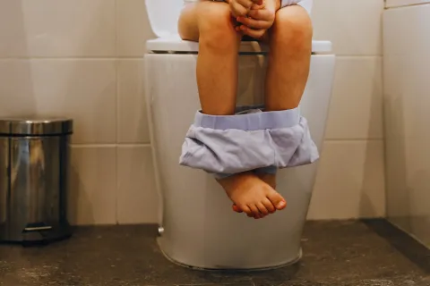 Child sitting on a toilet with pants down in a bathroom with tiled walls and floor.