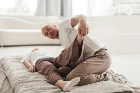 Man performing leg stretching exercise on woman lying on a mat in bright living room