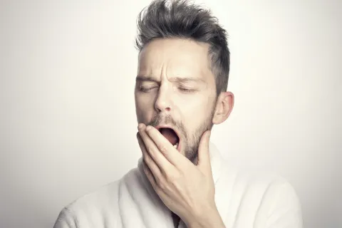 Young man with beard yawning and covering mouth, wearing a white robe against a neutral background