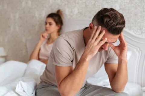 Stressed man sitting on bed holding his head with woman sitting behind looking upset, showing relationship tension.
