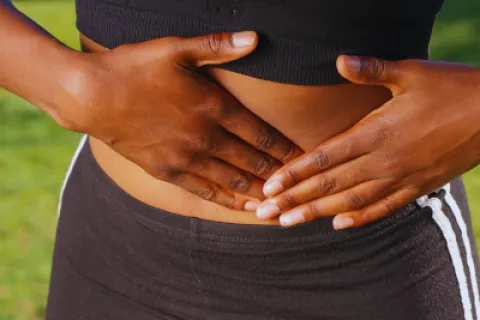 Woman wearing black workout clothes holding her stomach outdoors showing digestive discomfort.