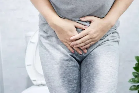 Woman in gray clothes clutching lower abdomen sitting on toilet, indicating urinary or abdominal discomfort.