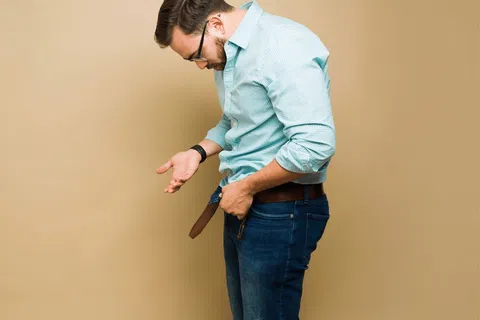Man in glasses adjusting his belt wearing a blue checkered shirt and jeans against a beige background