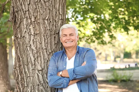 Smiling elderly man with white hair leans against a large tree trunk in a sunlit park with green foliage.