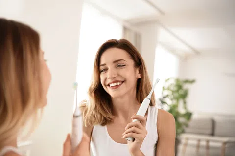 Smiling woman holding electric toothbrush in bathroom mirror, promoting dental hygiene and oral care routine.