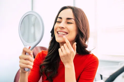 Young woman smiling and inspecting her teeth in a handheld mirror indoors with natural light.