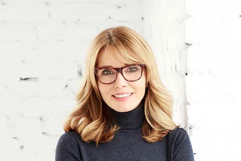 Smiling woman with blonde hair and glasses wearing a dark turtleneck sweater standing against a white brick wall.