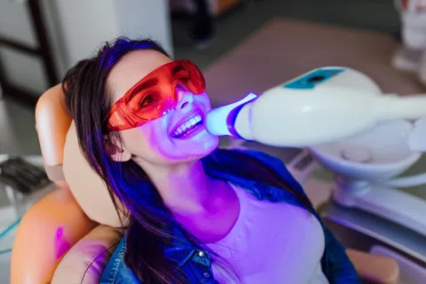 Woman wearing protective red glasses undergoing dental teeth whitening with a bright blue light device.