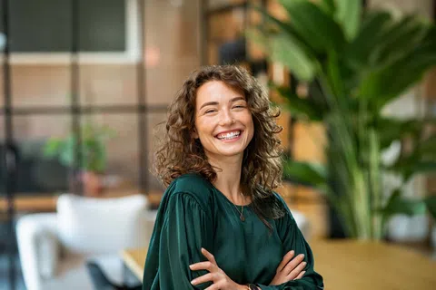 Smiling curly woman in green blouse standing with arms crossed in modern office with plants and blurred background