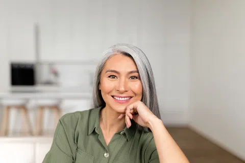 Smiling middle-aged woman with gray hair wearing green shirt in a bright modern kitchen.