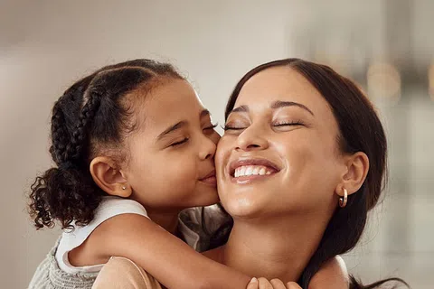 Young girl kisses her smiling mother on the cheek while hugging her tightly in a warm family moment.