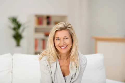 Smiling blonde woman sitting on white couch in a bright, modern living room with bookshelves and plants.