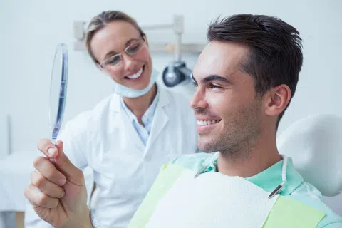 Smiling man checks teeth in handheld mirror while dentist observes in bright dental office