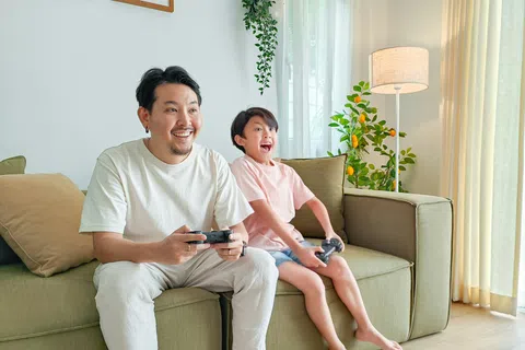 Father and son playing video games together on a sofa in a bright, cozy living room with plants and natural light.