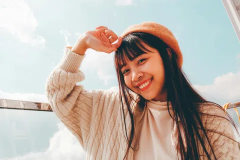 Young woman wearing an orange beret and sweater smiles outdoors under a blue sky with clouds