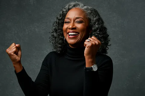 Joyful middle-aged woman with curly gray hair smiling and celebrating with raised fists against gray background