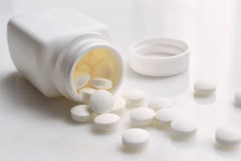 White tablets spilling from an open white plastic pill bottle on a light surface with the bottle cap beside it.