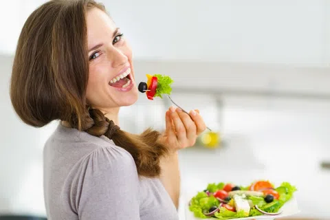 Woman with braided hair happily eating a fresh vegetable salad with olives and lettuce in a bright kitchen.