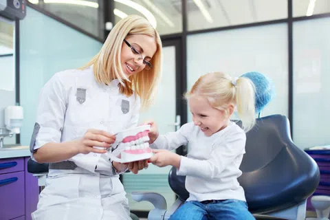 Female dentist teaching little girl about dental care using a teeth model in a bright clinic.