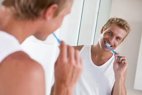 Young man in white tank top brushing his teeth in front of a bathroom mirror smiling.