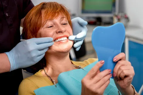 Woman smiling with healthy white teeth during dental checkup, holding a blue tooth-shaped mirror.