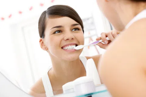 Young woman smiling while brushing her teeth in front of a bathroom mirror with dental products nearby